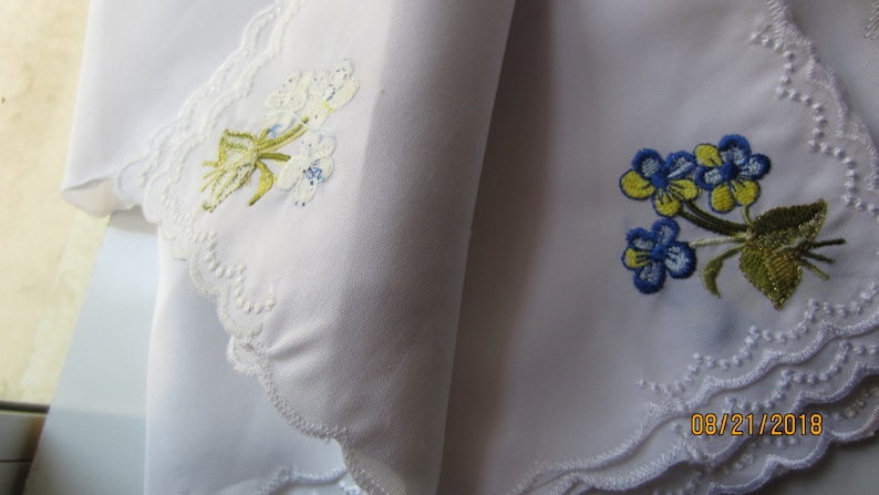 Puede incluir: Dos pa&ntilde;uelos blancos con dise&ntilde;os florales bordados. Un pa&ntilde;uelo presenta flores blancas con tallos y hojas verdes. El otro pa&ntilde;uelo presenta flores azules y amarillas con tallos y hojas verdes. Ambos pa&ntilde;uelos tienen un borde de encaje blanco.