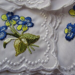 Puede incluir: Tela blanca con un delicado borde de encaje y flores azules y amarillas bordadas. La tela tiene una fecha bordada: 08/21/2018.