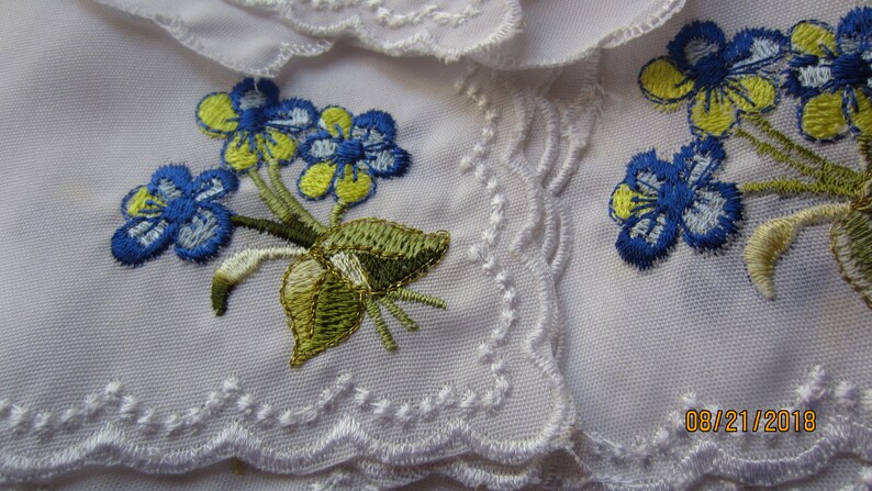Puede incluir: Tela blanca con un delicado borde de encaje y flores azules y amarillas bordadas. La tela tiene una fecha bordada: 08/21/2018.