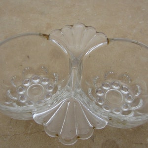 Puede incluir: Un plato doble de vidrio transparente con un asa decorativa. El plato tiene un borde festoneado y un diseño en relieve de pequeños círculos alrededor del centro de cada cuenco.