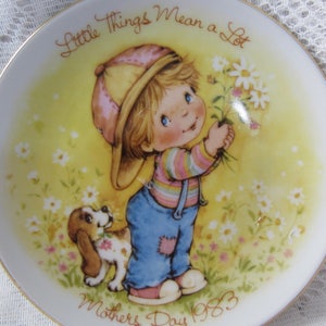 May include: A white ceramic plate with a gold rim features a colorful illustration of a child holding a bouquet of flowers. The text "Little Things Mean a Lot" is written in a cursive font above the child. The text "Mother's Day 1983" is written below the child.