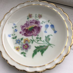 May include: A white porcelain dish with a gold rim and a floral design. The design features a large purple flower with yellow and green leaves, and smaller blue and yellow flowers.
