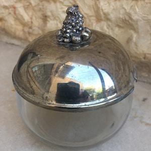 May include: A silver sugar bowl with a glass base and a grape-shaped lid. The lid is made of metal and has a detailed design of grapes.