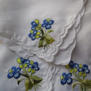 Puede incluir: Tres pa&ntilde;uelos de lino blanco con flores bordadas azules y amarillas. Los pa&ntilde;uelos tienen un borde festoneado.