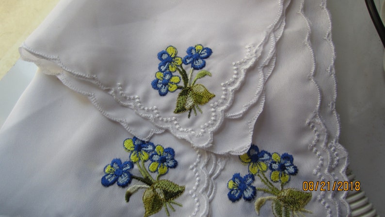 Puede incluir: Tres pa&ntilde;uelos de lino blanco con flores bordadas azules y amarillas. Los pa&ntilde;uelos tienen un borde festoneado.