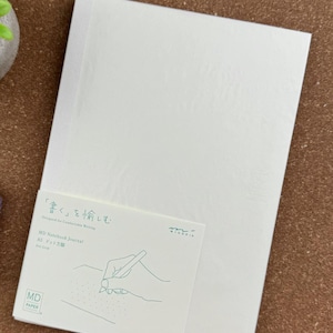 May include: A white MD Notebook Journal with a paper band featuring the text "書く」を愉しむ", "Designed for Comfortable Writing", "MD Notebook Journal", "A5 ドット方眼", "Dot Grid", "MIDORI", and "MD PAPER" alongside a line drawing of a hand writing.