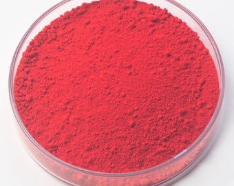 May include: A clear, circular petri dish containing a bright red powder. The powder has a fine, granular texture and completely fills the dish. The dish is set against a plain white background, highlighting the vivid colour of the powder.