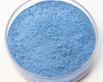 May include: A clear glass petri dish filled with a fine, light blue powder. The powder has a textured, granular appearance, and the dish is set against a plain white background. The image is a close-up, highlighting the color and texture of the powder.