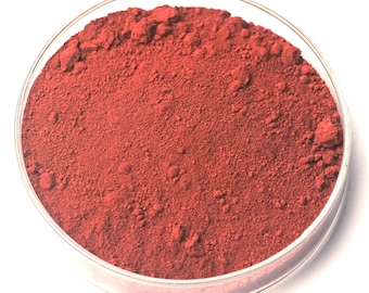 May include: A close-up view of a clear, round petri dish filled with a vibrant, reddish-brown powder. The powder has a fine, granular texture and fills the dish completely. The dish is set against a bright white background.