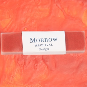 May include: A rectangular, red-orange block with the text "MORROW ARCHIVAL Realgar" on a white label. The block is set against a vibrant, textured orange background, creating a striking contrast. The product appears to be a type of art supply.
