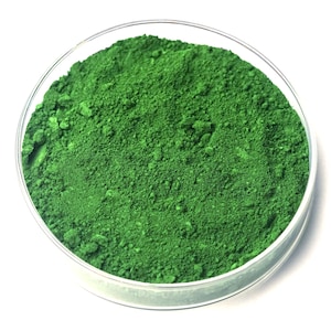 May include: A circular glass dish containing a vivid green powder. The powder appears finely ground and textured, possibly a pigment or dye. The dish is set against a bright white background.