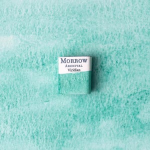 May include: A small, rectangular watercolour paint block with the text "MORROW ARCHIVAL Viridian" in white on a white and teal label. The paint block is teal and sits on a teal watercolour paper background.