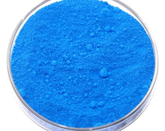 May include: A close-up shot of a clear glass petri dish filled with a vibrant, bright blue powder. The powder has a fine, granular texture and fills the dish completely. The dish is set against a plain white background.