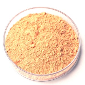May include: A clear glass petri dish filled with a light orange-brown powder. The powder has a fine, granular texture and fills the dish completely. The dish is set against a white background.