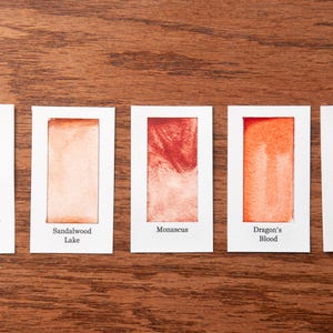 five small white cards with different colored paint swatches on them, arranged on a wooden surface.