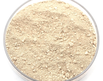 Pode incluir: Uma placa de Petri de vidro transparente preenchida com um pó granular bege claro. O pó tem uma aparência fina e texturizada com pequenos aglomerados irregulares. A placa está sobre um fundo branco brilhante.