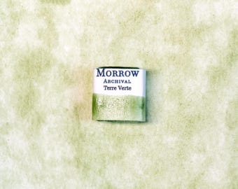 May include: A small, rectangular watercolor paint block with the text "MORROW ARCHIVAL Terre Verte" in a white and green color scheme. The paint block is set against a textured, light green background, suggesting a focus on art supplies.