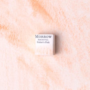 Puede incluir: Un pequeño bloque de pintura de acuarela cuadrado, etiquetado como "MORROW ARCHIVAL Potter's Pink", en un tono rosa claro y suave. El fondo es un lavado de acuarela texturizado en un tono rosa similar.