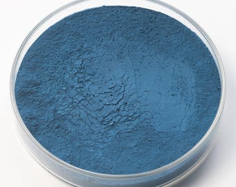 May include: A clear, round petri dish filled with a fine, textured blue powder. The powder has a slightly uneven surface, with visible ridges and crevices. The dish is set against a plain white background.
