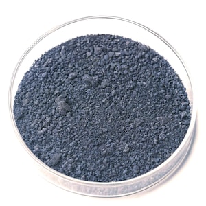 Op de afbeelding: Een heldere glazen petrischaal gevuld met een donkerblauwe korrelige substantie. De substantie is een fijn poeder of kleine deeltjes, met een enigszins ongelijke textuur. De schaal staat tegen een heldere witte achtergrond.