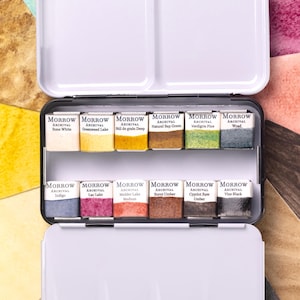 Könnte beinhalten: Eine offene, weiße Metall-Aquarellfarbpalette mit zwei Fächern und zehn rechteckigen Farbnäpfchen. Die Näpfchen sind mit verschiedenen Farben beschriftet, darunter "Bone White", "Indigo" und "Burnt Umber".