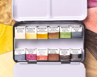 May include: A white metal watercolor paint palette with two sections and ten rectangular paint pans. The pans are labeled with various colors, including "Bone White," "Indigo," and "Burnt Umber." The palette is open, revealing the paints.