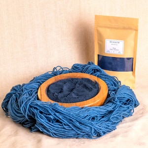 Op de afbeelding: Een houten kom gevuld met donkerblauw poeder ligt op een stapel blauw garen. Een bruine papieren zak met een etiket en een venster met donkerblauw poeder staat op de achtergrond. Het etiket luidt "Morrow Botanical".