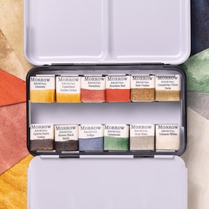Könnte beinhalten: Geöffnete, weiße Metall-Aquarellfarbpalette mit zwei Mischbereichen. Die Palette enthält zwölf rechteckige Farbtöpfchen mit verschiedenen Farben, die jeweils mit der Marke "MORROW" und Farbbezeichnungen wie "Limonite" und "Vermilion" versehen sind.