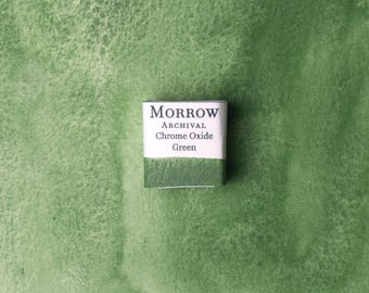 May include: A square watercolor paint pan labeled "MORROW ARCHIVAL Chrome Oxide Green" sits on a textured green surface. The paint pan is white with black text and a green paint swatch.