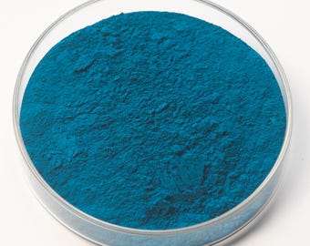 May include: A clear, round petri dish filled with a vibrant, teal-colored powder. The powder has a fine, textured appearance, and the dish is set against a plain white background. The image is a close-up, highlighting the color and texture of the powder.