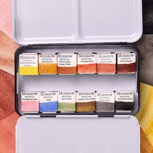 May include: A metal watercolor paint set with a white lid and a black base. The set contains twelve rectangular paint pans with various colors, including yellow, orange, red, and green. The brand name "MORROW ARCHIVAL" is printed on each pan.