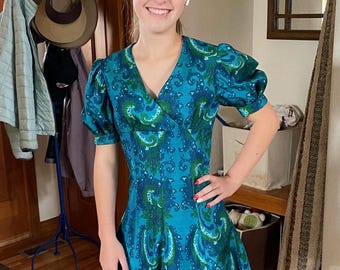 May include: A teal and green paisley print dress with short, puffed sleeves and a V-neck. The dress has a wrap-style bodice and a flared skirt. The dress is knee-length and has a retro, vintage style.