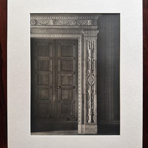 May include: A black and white photographic print matted in white, depicting a set of double doors with recessed panels and a highly detailed, carved stone door frame featuring intricate floral and geometric motifs, presented on a dark wood surface.
