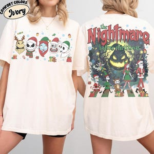 Op de afbeelding: Een ivoorkleurig t-shirt met The Nightmare Before Christmas-afbeeldingen. De voorkant toont karakterhoofden met kerstmutsen, terwijl de achterkant een grote scène toont met personages, de tekst The Nightmare before christmas en een volle maan.