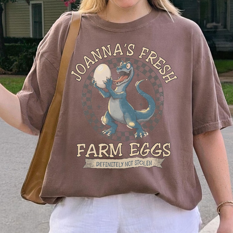 May include: A brown t-shirt featuring a cartoon dinosaur holding an egg, with the text "JOANNA'S FRESH FARM EGGS" and "DEFINITELY NOT STOLEN". The design is centered on the shirt.