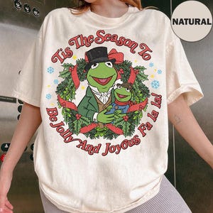 Puede incluir: Camiseta color crema con Kermit la Rana y un personaje de rana más pequeño en una corona navideña. El texto dice "Tis The Season To Be Jolly And Joyous Fa la la!" en una fuente festiva. Una etiqueta "NATURAL" es visible.