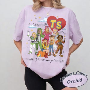 May include: An orchid-colored t-shirt featuring a graphic of seven characters dressed in various costumes. A red heart with the yellow text TS sits above them. Musical notes, clouds, and the text I knew it, I knew you are printed below.