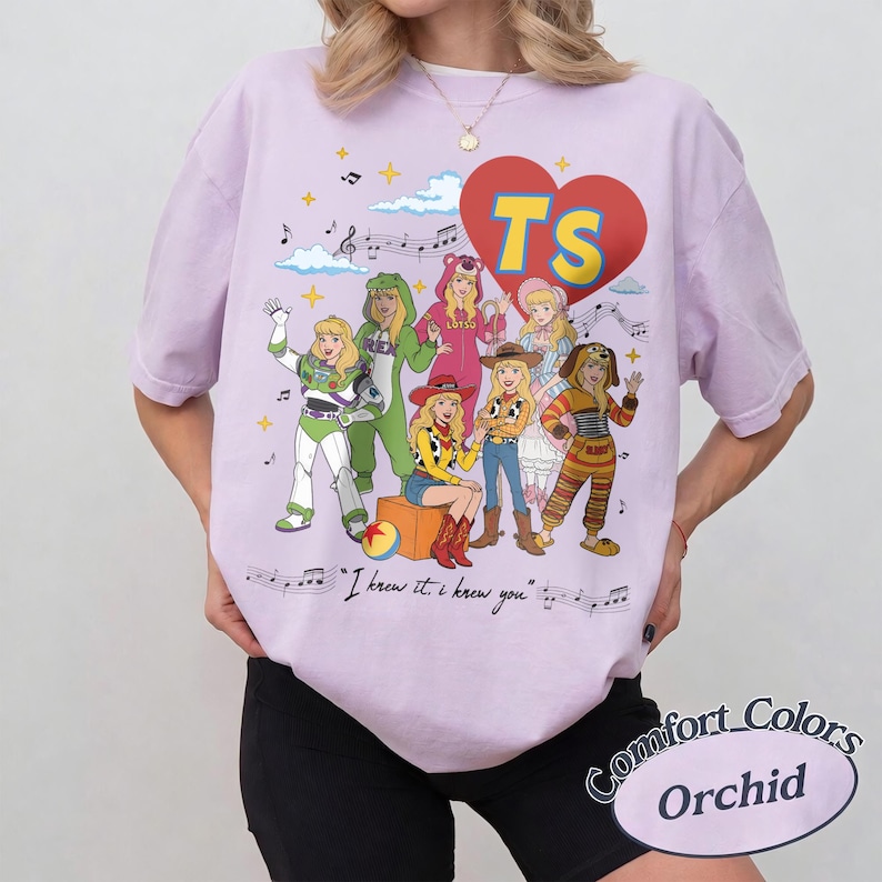 May include: An orchid-colored t-shirt featuring a graphic of seven characters dressed in various costumes. A red heart with the yellow text TS sits above them. Musical notes, clouds, and the text I knew it, I knew you are printed below.
