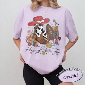 Pu&ograve; includere: Una t-shirt viola chiaro con una stampa grafica di un cappello da cowboy rosso, uno stivale marrone e bianco con stampa mucca, una chitarra acustica, blocchi dell'alfabeto e un microfono. Il testo recita, I knew it, i knew you. Comfort Colors Orchid.