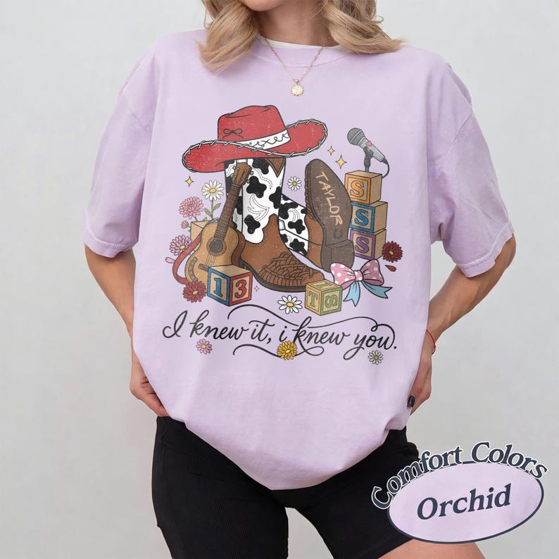 Pu&ograve; includere: Una t-shirt viola chiaro con una stampa grafica di un cappello da cowboy rosso, uno stivale marrone e bianco con stampa mucca, una chitarra acustica, blocchi dell'alfabeto e un microfono. Il testo recita, I knew it, i knew you. Comfort Colors Orchid.