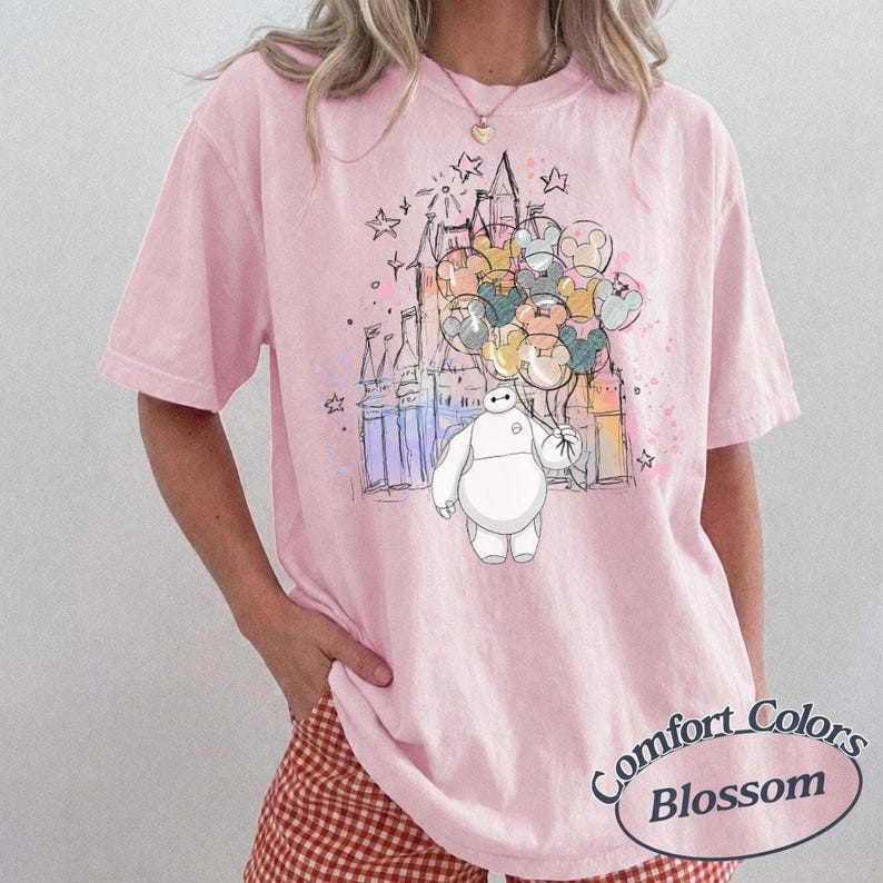 Puede incluir: Camiseta rosa p&aacute;lido Comfort Colors Blossom con una ilustraci&oacute;n de dibujos animados de Baymax sosteniendo globos frente a un castillo. El dise&ntilde;o incluye estrellas y un efecto acuarela. La camiseta tiene mangas cortas.