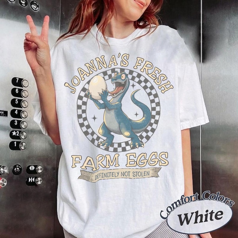 May include: White t-shirt with a dinosaur graphic holding an egg, surrounded by text that reads "Joanna's Fresh Farm Eggs, Definitely Not Stolen." The shirt also has "Comfort Colors" and "White" printed on it.