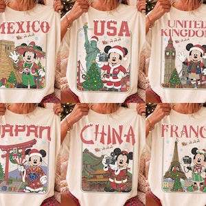 May include: A cream-colored t-shirt featuring six rectangular graphic panels. Each panel displays Mickey Mouse in a different country—Mexico, USA, United Kingdom, Japan, China, and France—wearing festive holiday attire and iconic landmarks.