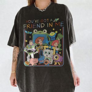 May include: A charcoal gray t-shirt featuring a colorful graphic print of various animated characters from the Toy Story franchise. The text "YOU'VE GOT A FRIEND IN ME" is printed in a multi-colored, distressed font above the character illustration.