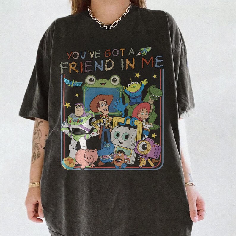 May include: A charcoal gray t-shirt featuring a colorful graphic print of various animated characters from the Toy Story franchise. The text "YOU'VE GOT A FRIEND IN ME" is printed in a multi-colored, distressed font above the character illustration.