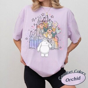 Puede incluir: Camiseta color orqu&iacute;dea con un gr&aacute;fico de Baymax sosteniendo globos de Mickey Mouse frente a un castillo. El dise&ntilde;o incluye estrellas y detalles de estilo acuarela. La camiseta tiene mangas cortas y un corte holgado.