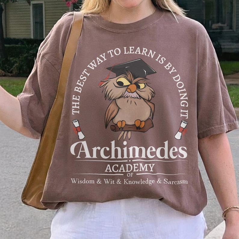 May include: A brown t-shirt featuring a graphic of an owl wearing a graduation cap, perched on a branch. The text reads: THE BEST WAY TO LEARN IS BY DOING IT, Archimedes ACADEMY OF WISDOM & WIT & KNOWLEDGE & SARCASM, with two diploma scrolls.