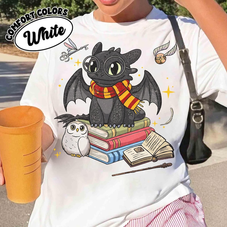 May include: White t-shirt featuring a cartoon dragon wearing glasses and a scarf, sitting on books. An owl, a golden snitch, and other magical elements are also depicted. The text "COMFORT COLORS White" is in a white oval.