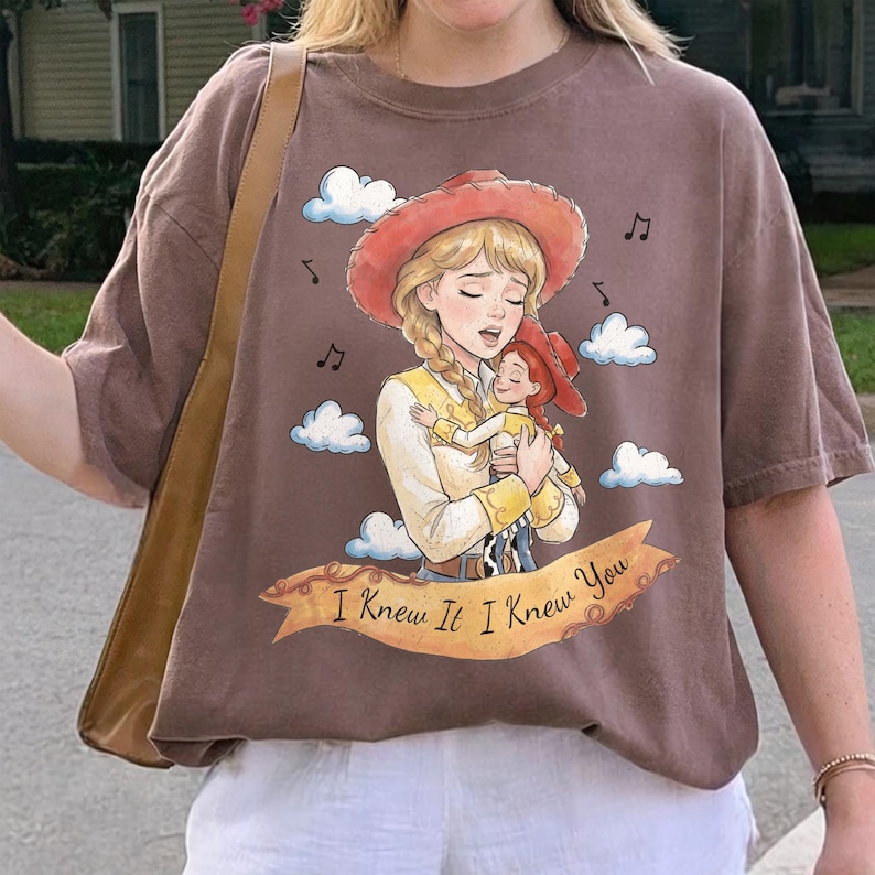 May include: A brown short-sleeve t-shirt featuring a graphic illustration of a blonde cowgirl holding a smaller doll. The design includes white clouds, musical notes, and a tan banner with the text I Knew It I Knew You printed in black.