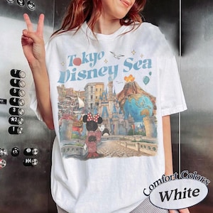 Pu&ograve; includere: T-shirt bianca con grafica "Tokyo Disney Sea" in testo blu e rosso. Il design include illustrazioni di un castello, Minnie Mouse e un globo. La maglietta &egrave; etichettata "Comfort Colors White".
